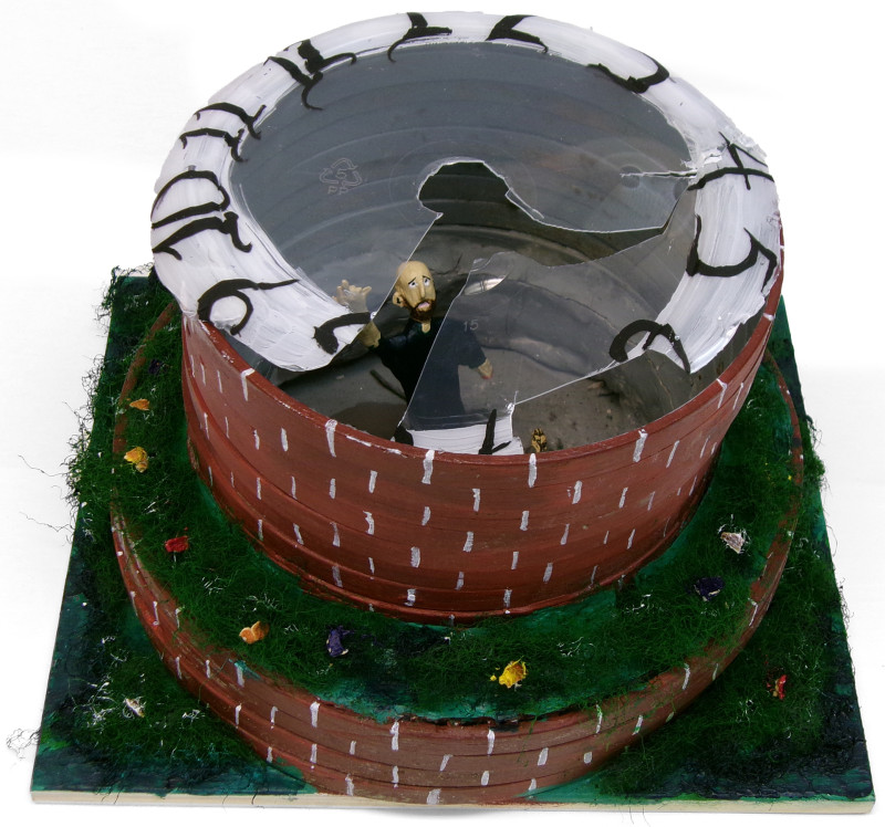 A sculpture of a physical enclosure and a small person. The enclosure is made of two circular tiers. The walls of the tiers are reddish brown with white details. The ground, and the horizontal area above the first tier, is covered in grass and what look like flowers. At the top, there is a piece of clear, cracked plastic. The edges of the plastic are painted white, with black numerals representing the face of a clock. A bald man with a beard is within the structure. He has one arm lifted and he looks toward the viewer.