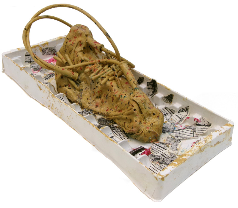 An abstract sculpture of what might be a bodily organ, a futuristic shoe, or just a shape. The object is contained in a white plastic tray that acts as pedestal. There are bits of paper scattered across the surface of the tray. The shape itself is yellow-brown, and multicolor paint splatters appear throughout the entire sculpture. Shapes like tubes, bullet casings, or pasta emerge from the top of the main object.