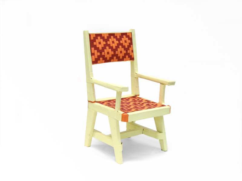 A sculpture of a chair made of folded paper. The structural elements are cream, the seat and seat back are woven to create a diamond pattern, from light orange and red.