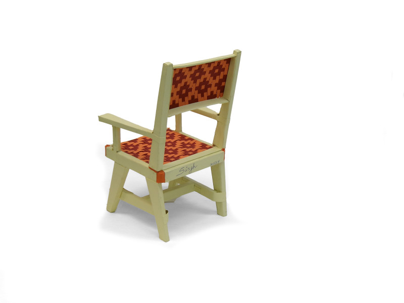 A sculpture of a chair made of folded paper, seen from the back. The structural elements are cream, the seat and seat back are woven to create a diamond pattern, from light orange and red.
