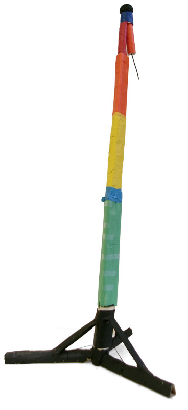 A tall, skinny structure like a tripod. A central column is wrapped in a color-blocked rainbow of paper, with green at the bottom, a little bit of blue, then yellow, then orange, with red on top. At its highest point, the column looks folded over, with a tip dangling downwards and a string hanging further than its end. There is blue tape at this breaking point.